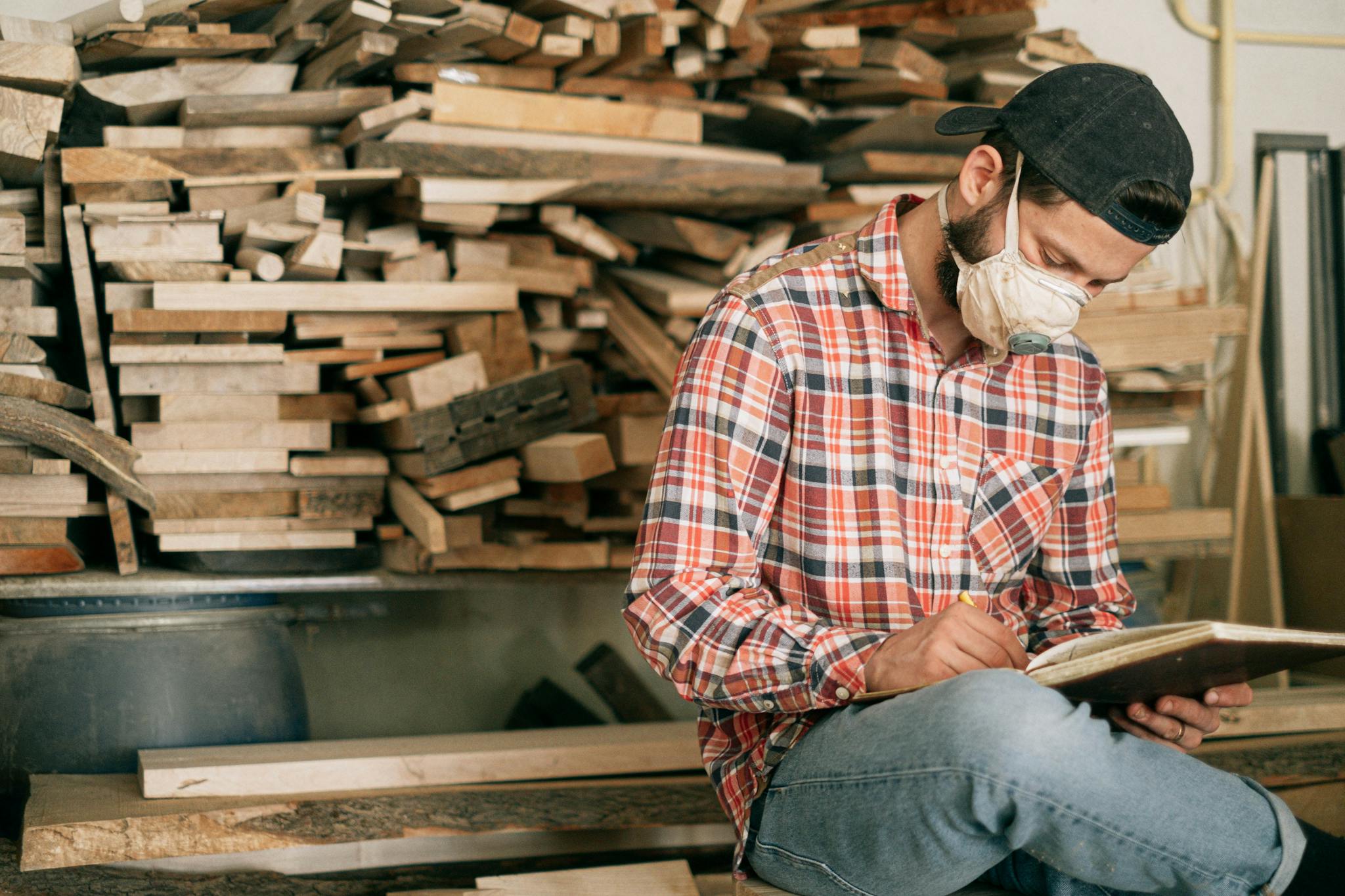 A carpenter in a mask designs plans amidst stacks of wood in an indoor workshop.